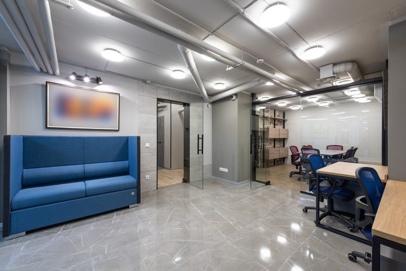 A modern office lobby with a blue couch, glass-walled meeting room, and polished tile floor