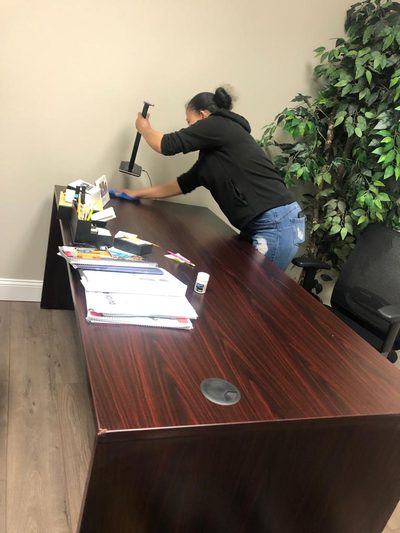 A KI Cleaning staff member wiping down a wood office desk beside a potted plant