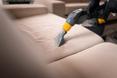 A gloved hand using an upholstery attachment to clean a beige fabric sofa cushion