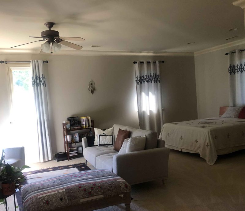 A tidy home bedroom and sitting area with a made bed, gray sofa, and curtained windows