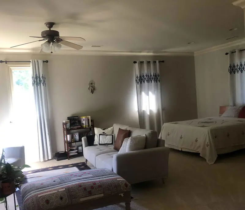 A tidy home bedroom and sitting area with a made bed, gray sofa, and curtained windows
